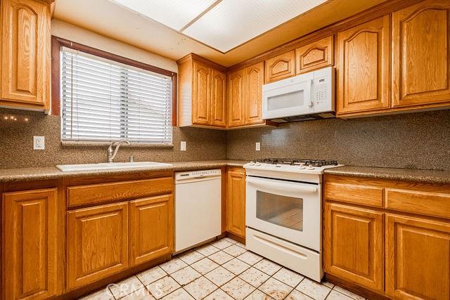 kitchen with brown cabinetry, backsplash, a sink, light tile patterned floors, and white appliances