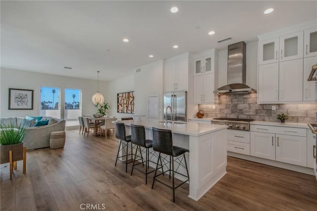 kitchen with wall chimney range hood, a kitchen island with sink, light countertops, stainless steel appliances, and dark wood-type flooring