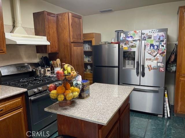 kitchen featuring brown cabinets, stainless steel fridge, gas stove, wall chimney exhaust hood, and freestanding refrigerator