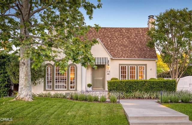 tudor house with a chimney, roof with shingles, stucco siding, and a front yard