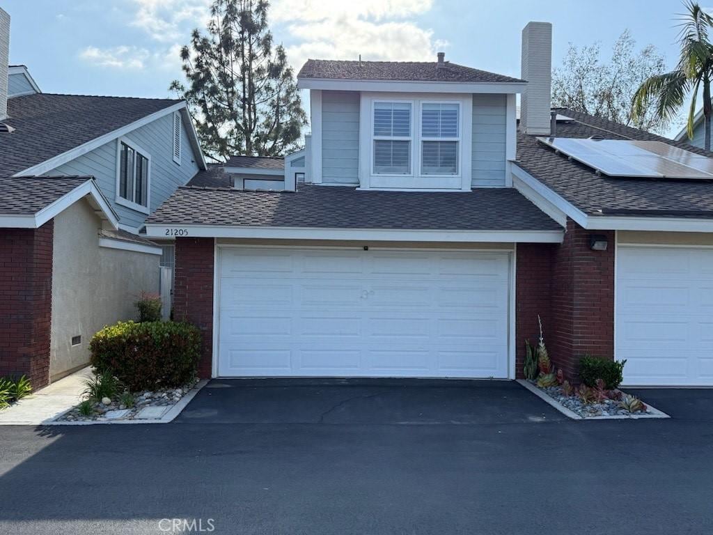view of front facade with driveway, a chimney, roof with shingles, brick siding, and roof mounted solar panels