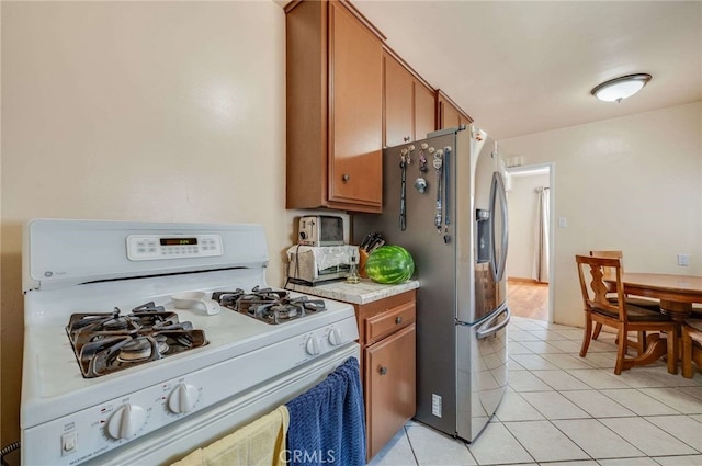 kitchen featuring brown cabinets, light countertops, light tile patterned floors, stainless steel refrigerator with ice dispenser, and white range with gas cooktop