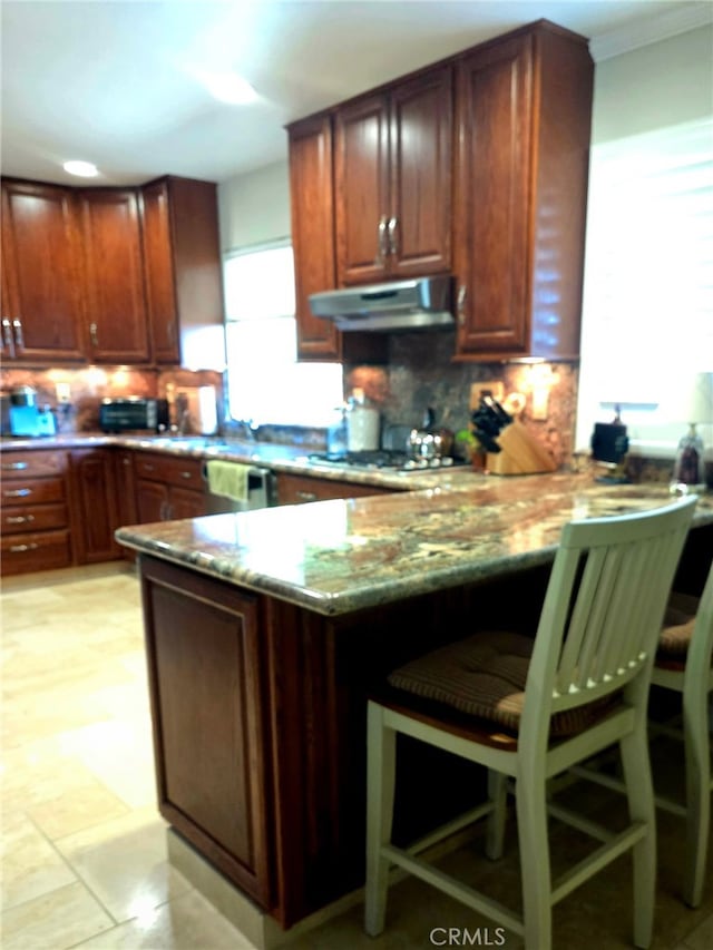 kitchen with crown molding, decorative backsplash, a peninsula, a breakfast bar, and under cabinet range hood