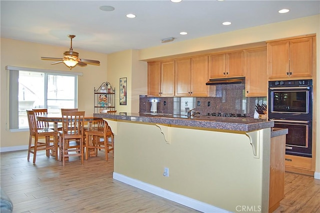 kitchen with dobule oven black, a breakfast bar area, under cabinet range hood, a ceiling fan, and backsplash