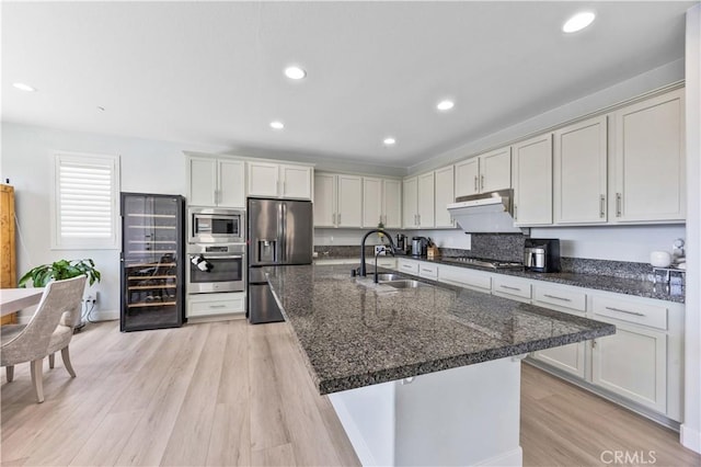 kitchen with beverage cooler, a center island with sink, light wood-type flooring, a sink, and stainless steel appliances