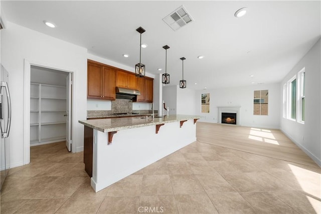 kitchen with under cabinet range hood, a lit fireplace, a breakfast bar area, brown cabinets, and open floor plan