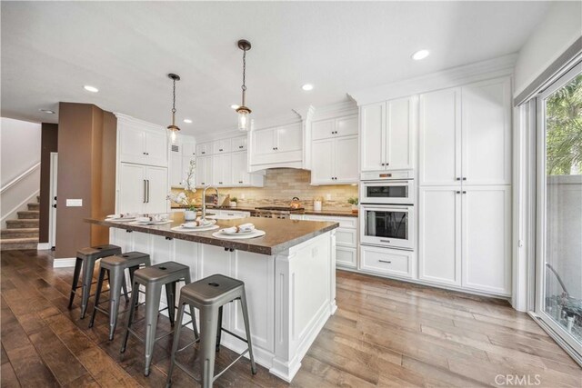 kitchen with wood-type flooring, paneled built in refrigerator, a kitchen island with sink, dark countertops, and white cabinetry
