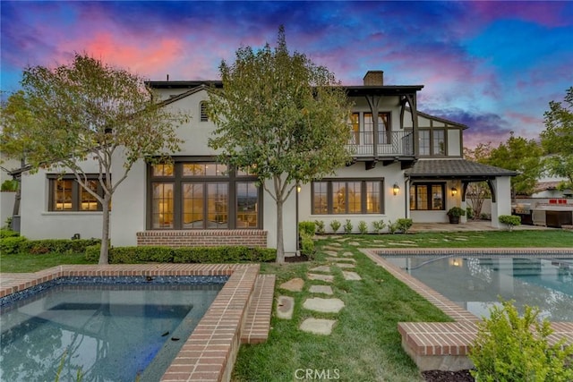 back of property featuring a balcony, an outdoor pool, stucco siding, a chimney, and a lawn