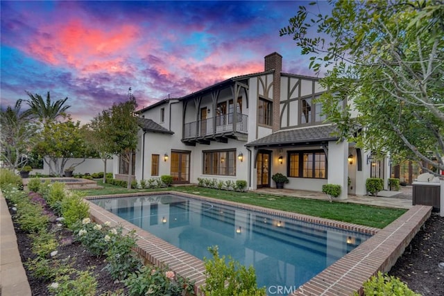 back of house at dusk featuring a balcony, an outdoor pool, stucco siding, a chimney, and a lawn