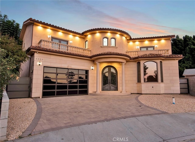 mediterranean / spanish-style house featuring a garage, a balcony, decorative driveway, stucco siding, and french doors