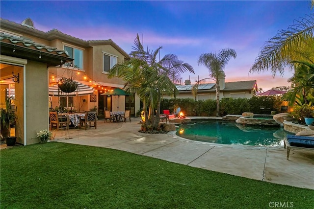 pool at dusk featuring a fire pit, a patio area, a pool with connected hot tub, and a yard