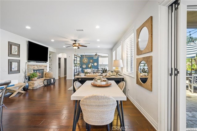 dining room with arched walkways, dark wood finished floors, a stone fireplace, a ceiling fan, and visible vents