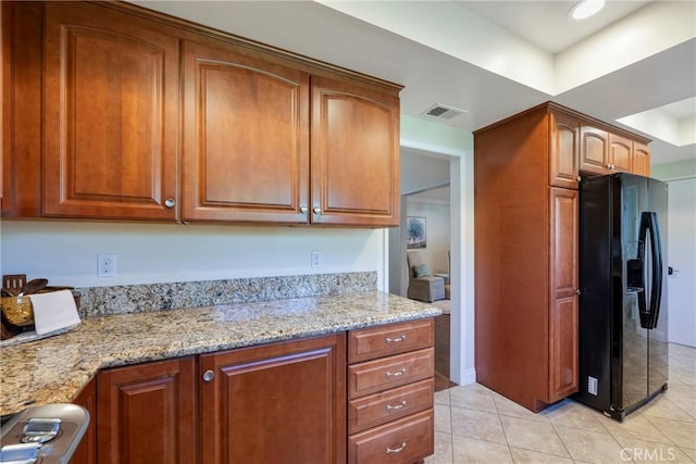 kitchen with light tile patterned flooring, light stone countertops, brown cabinetry, visible vents, and black fridge with ice dispenser
