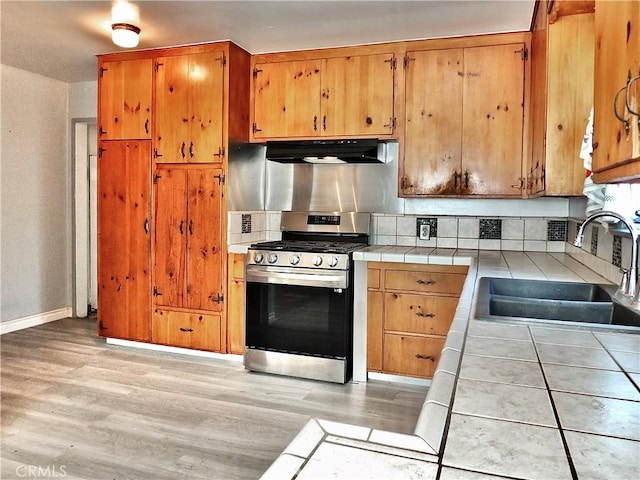 kitchen featuring stainless steel gas stove, light wood-style floors, tile counters, under cabinet range hood, and a sink