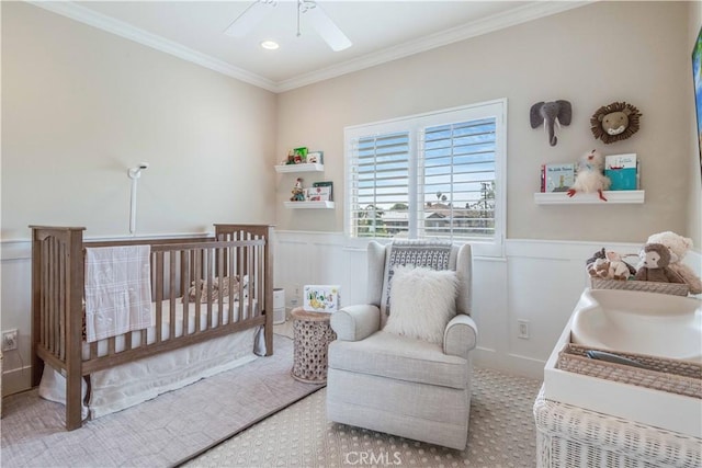 bedroom featuring ornamental molding, wainscoting, a ceiling fan, and a nursery area