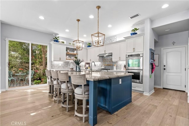 kitchen featuring a center island with sink, backsplash, stainless steel double oven, and white cabinetry