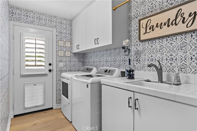 clothes washing area with cabinet space, a sink, wallpapered walls, light wood-type flooring, and washing machine and dryer