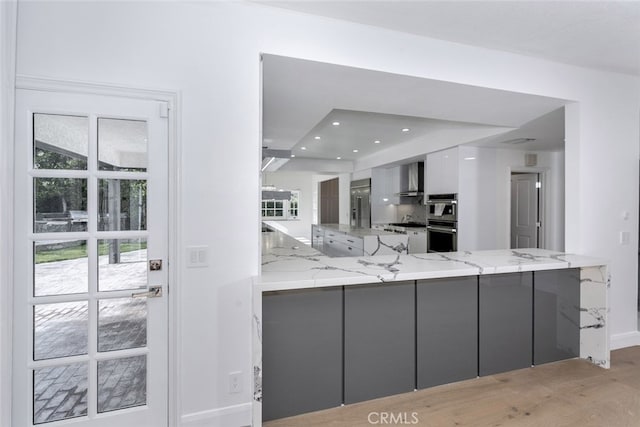 kitchen with wall chimney range hood, light stone countertops, double oven, gray cabinetry, and modern cabinets