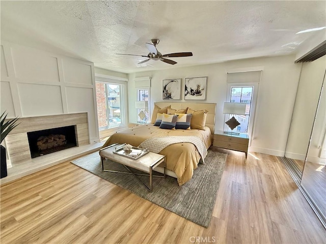 bedroom with a fireplace with raised hearth, a textured ceiling, light wood-style flooring, a ceiling fan, and baseboards