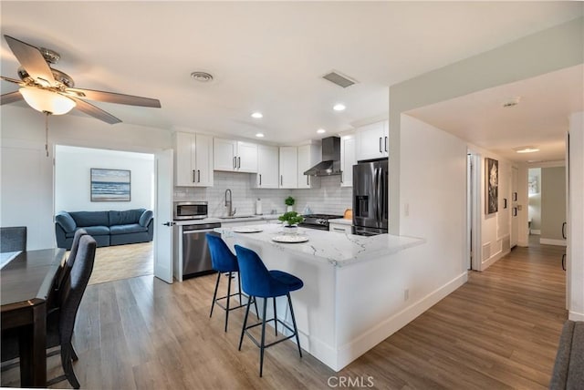 kitchen with wall chimney range hood, white cabinets, appliances with stainless steel finishes, a sink, and a kitchen bar
