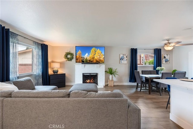 living area featuring baseboards, a brick fireplace, wood finished floors, and ceiling fan