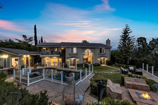 back of house at dusk featuring a balcony, a patio, an outdoor pool, stucco siding, and a chimney