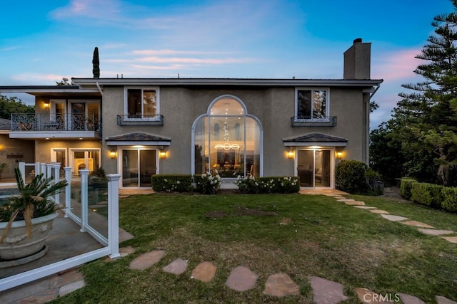 back of property at dusk featuring a lawn, stucco siding, a chimney, and a balcony