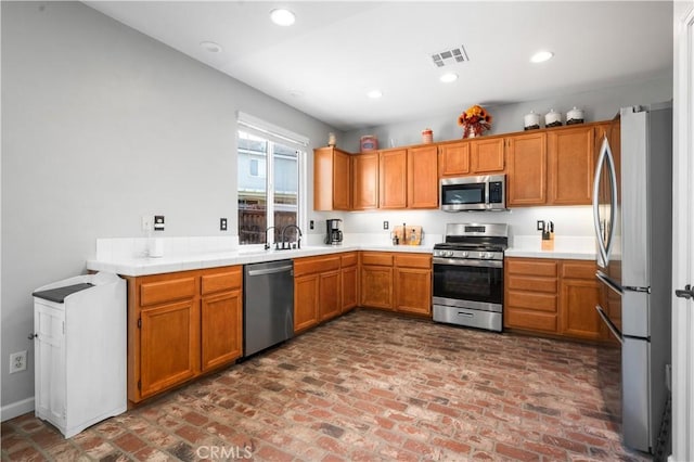kitchen with appliances with stainless steel finishes, brick floor, brown cabinetry, recessed lighting, and visible vents