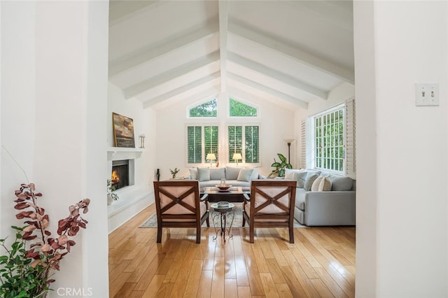living room featuring vaulted ceiling with beams, light wood-style floors, and a warm lit fireplace