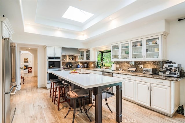 kitchen featuring a tray ceiling, a kitchen breakfast bar, tasteful backsplash, stainless steel appliances, and wall chimney exhaust hood