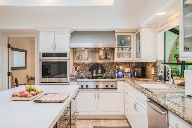 kitchen with white cabinets, appliances with stainless steel finishes, a sink, wall chimney exhaust hood, and backsplash