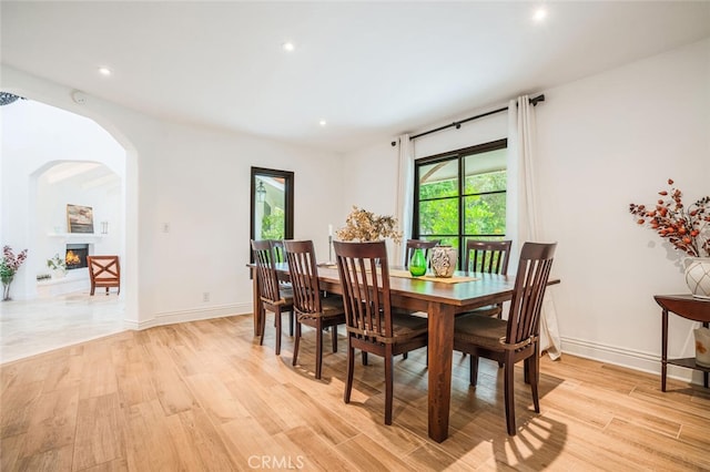 dining area featuring light wood-style floors, baseboards, a warm lit fireplace, recessed lighting, and arched walkways