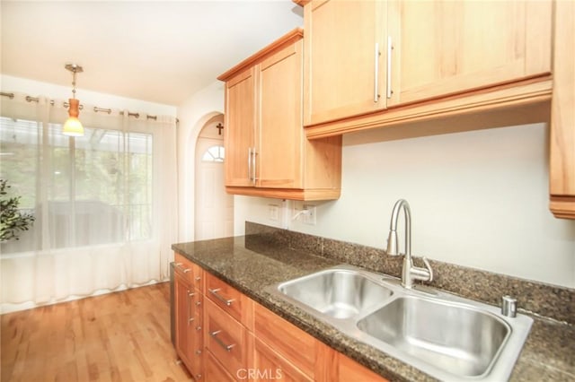 kitchen with arched walkways, hanging light fixtures, a sink, dark stone countertops, and light wood finished floors