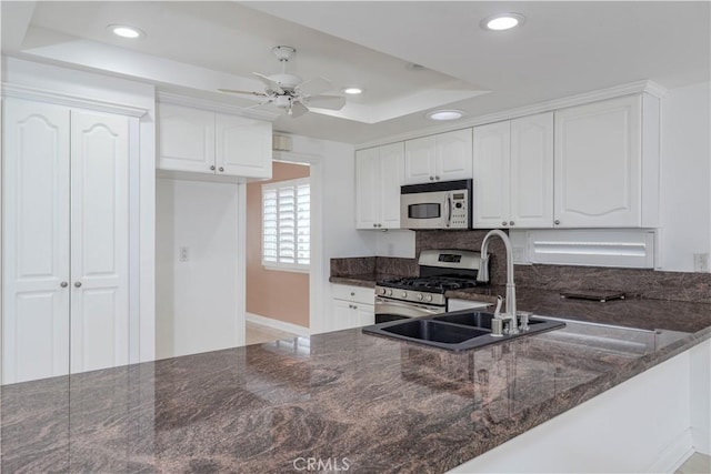 kitchen with a raised ceiling, stainless steel range with gas stovetop, a sink, dark stone counters, and ceiling fan