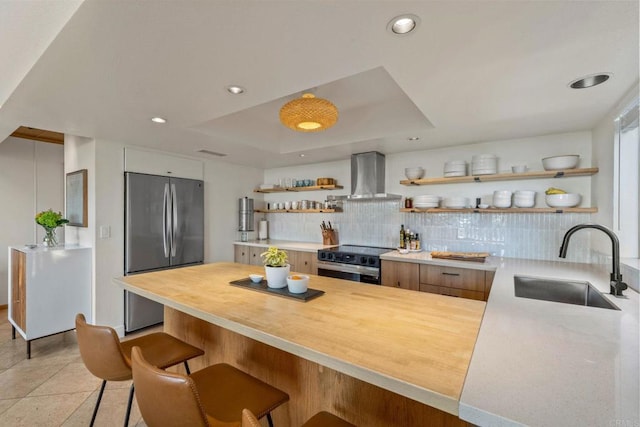 kitchen with open shelves, a peninsula, wall chimney range hood, a sink, and appliances with stainless steel finishes