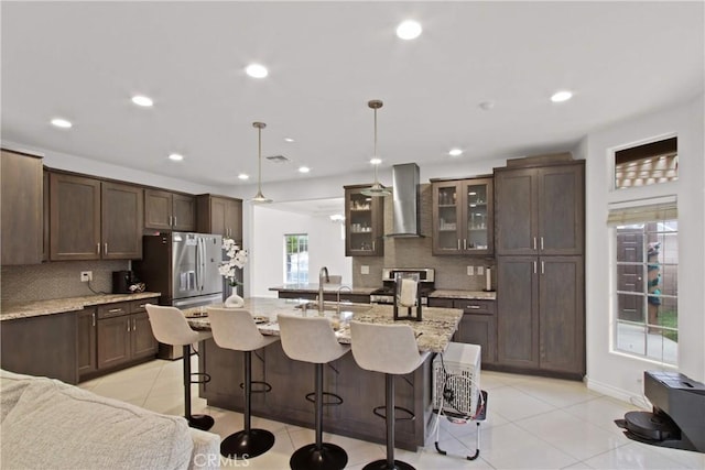 kitchen featuring wall chimney range hood, a kitchen breakfast bar, appliances with stainless steel finishes, tasteful backsplash, and light tile patterned floors