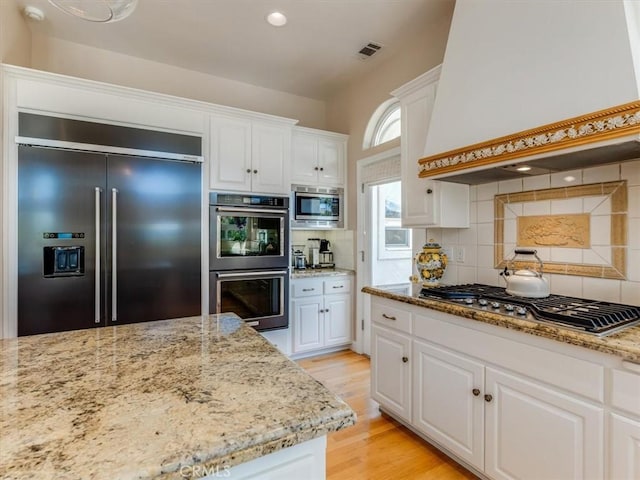 kitchen featuring light stone counters, white cabinetry, built in appliances, and premium range hood