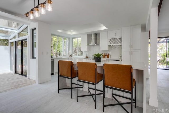 kitchen featuring wall chimney range hood, stainless steel dishwasher, light countertops, a raised ceiling, and white cabinetry