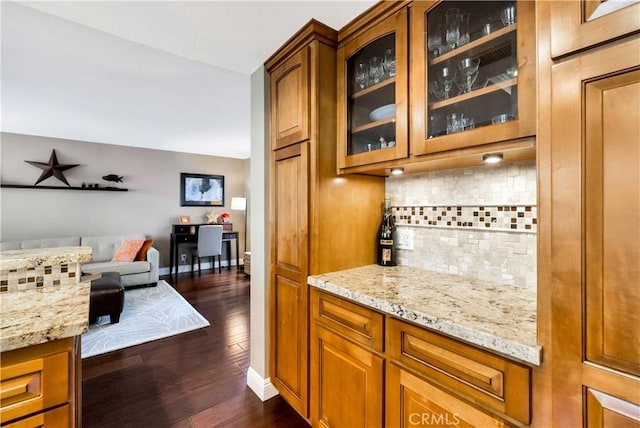 kitchen featuring dark wood-style flooring, light stone countertops, brown cabinetry, tasteful backsplash, and glass insert cabinets
