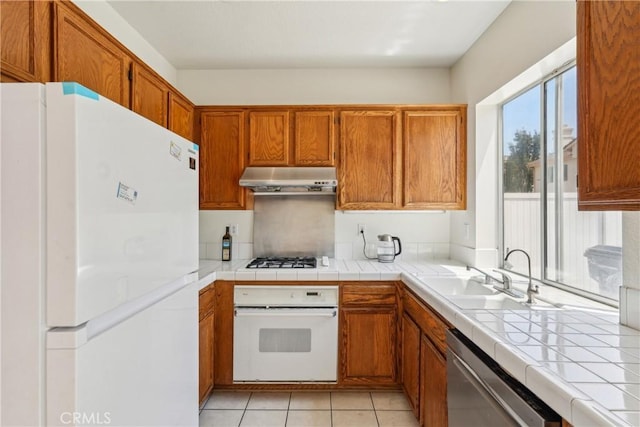 kitchen with brown cabinets, a sink, white appliances, light tile patterned floors, and under cabinet range hood