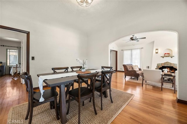 dining room featuring light wood-style floors, arched walkways, a ceiling fan, and a lit fireplace