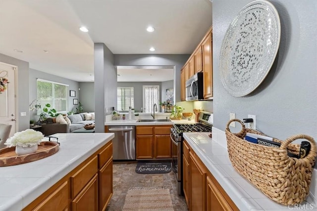 kitchen with a sink, brown cabinetry, stainless steel appliances, tile countertops, and a wealth of natural light