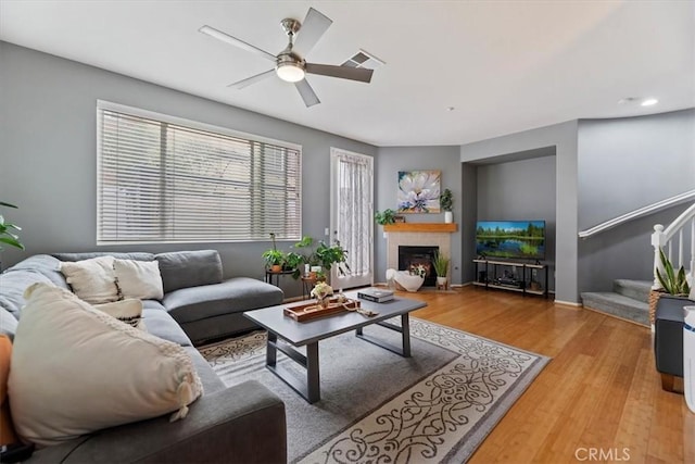 living room with ceiling fan, visible vents, a warm lit fireplace, light wood finished floors, and stairs