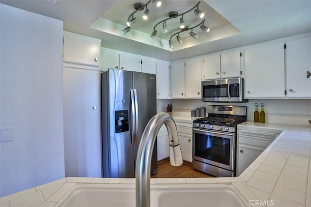 kitchen featuring appliances with stainless steel finishes, a sink, a raised ceiling, tile countertops, and white cabinetry