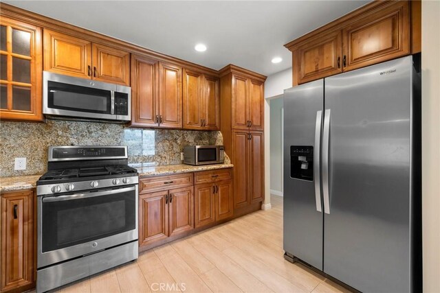 kitchen with brown cabinets, appliances with stainless steel finishes, light stone counters, light wood finished floors, and decorative backsplash