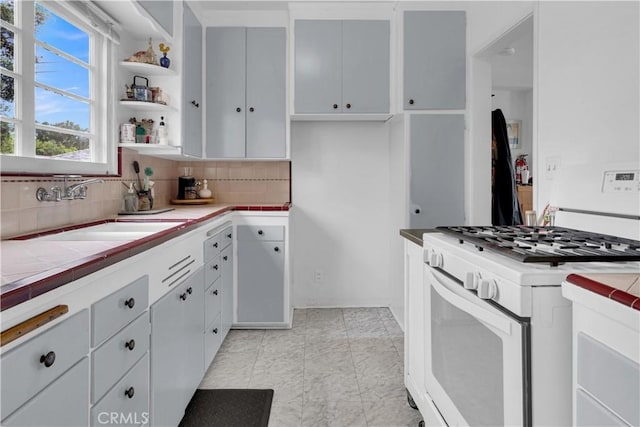 kitchen with decorative backsplash, a sink, open shelves, tile counters, and white gas range oven