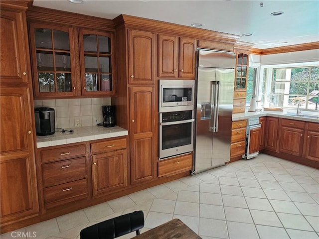 kitchen with a sink, built in appliances, brown cabinetry, and glass insert cabinets