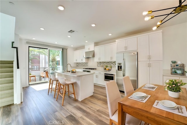 kitchen featuring stainless steel fridge with ice dispenser, light countertops, an island with sink, under cabinet range hood, and visible vents