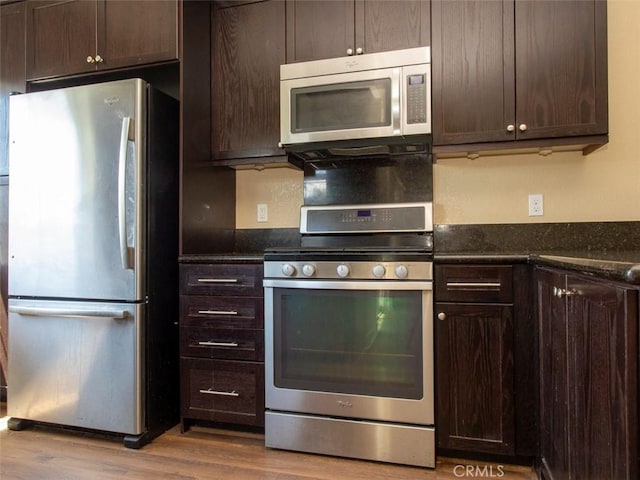 kitchen featuring stainless steel appliances, dark brown cabinetry, wood finished floors, and dark stone countertops