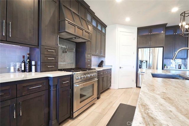 kitchen featuring dark brown cabinetry, appliances with stainless steel finishes, a sink, premium range hood, and backsplash
